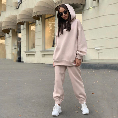 Frau in beigem Hoodie und Jogginghose, Sonnenbrille, weiße Sneaker, urbaner Hintergrund.