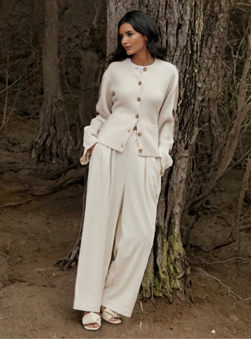 Frau in cremefarbenem Button-Up-Cardigan und Hose, lässig im Wald stehend.