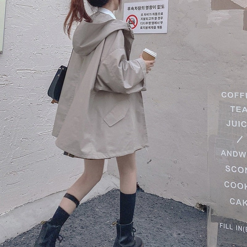 Frau in beigem Oversize-Mantel mit Kapuze, hält Kaffeebecher, trägt schwarze Stiefel.