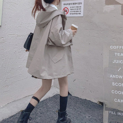 Frau in beigem Oversize-Mantel mit Kapuze, hält Kaffeebecher, trägt schwarze Stiefel.