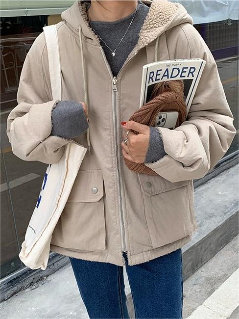 Frau in beiger Kapuzenjacke mit Buch und Handy, lässiger Herbstlook, Straßenhintergrund.