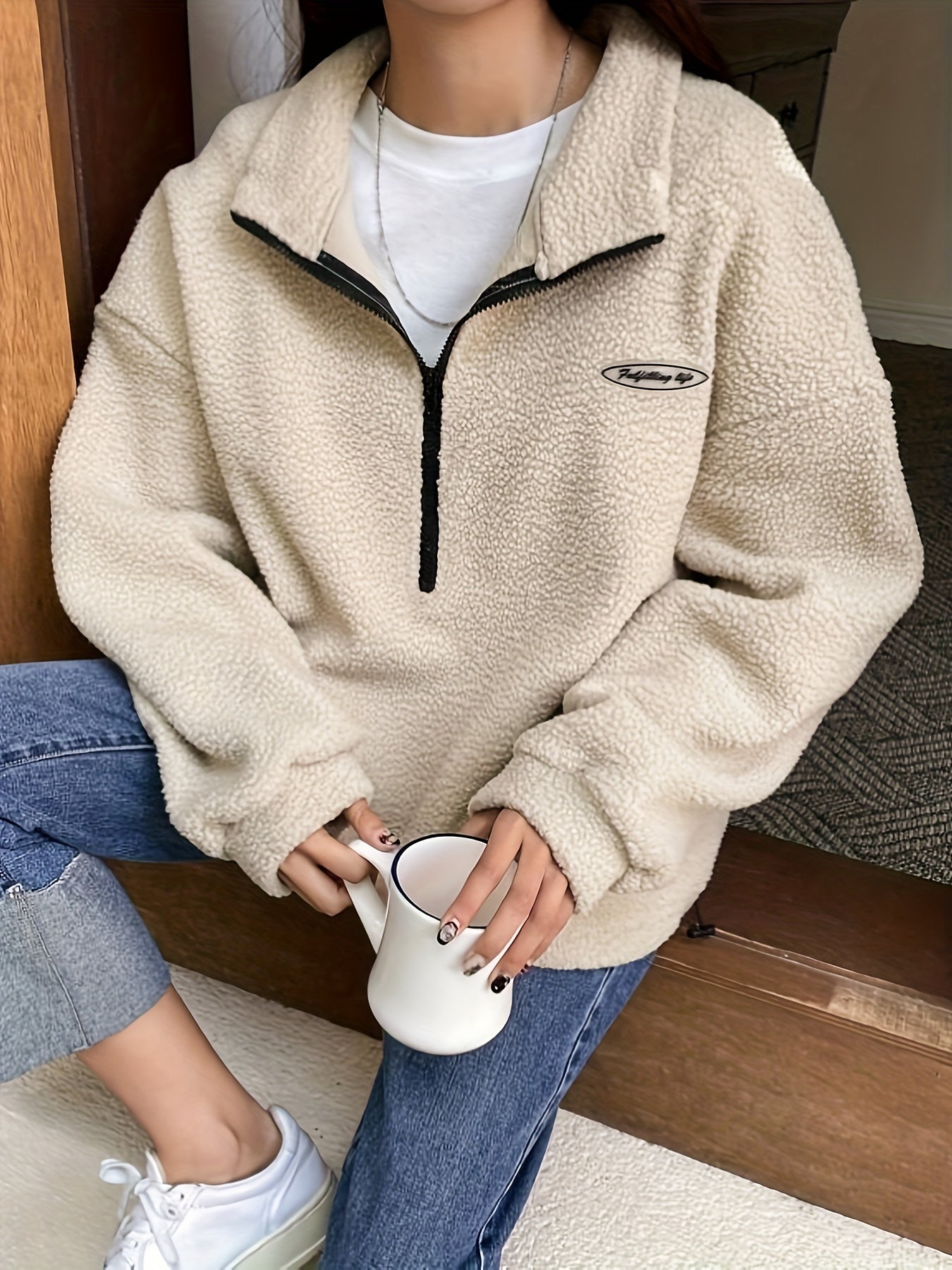 Frau in beigem Fleece-Pullover mit Reißverschluss, hält weiße Tasse, lässige Mode.