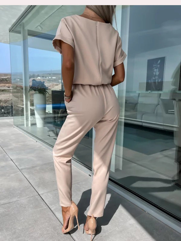 Frau in beigem Jumpsuit und High Heels auf moderner Terrasse mit Glaswänden.
