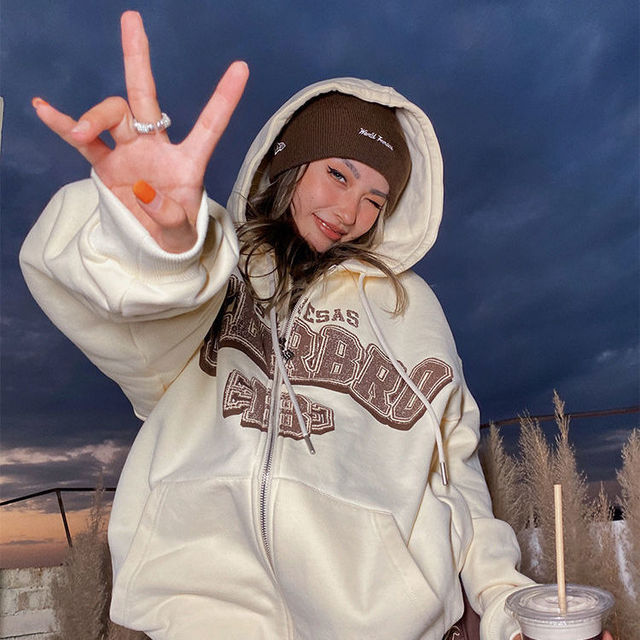 Frau in cremefarbenem Hoodie und brauner Mütze, lächelnd, bei Sonnenuntergang im Freien.