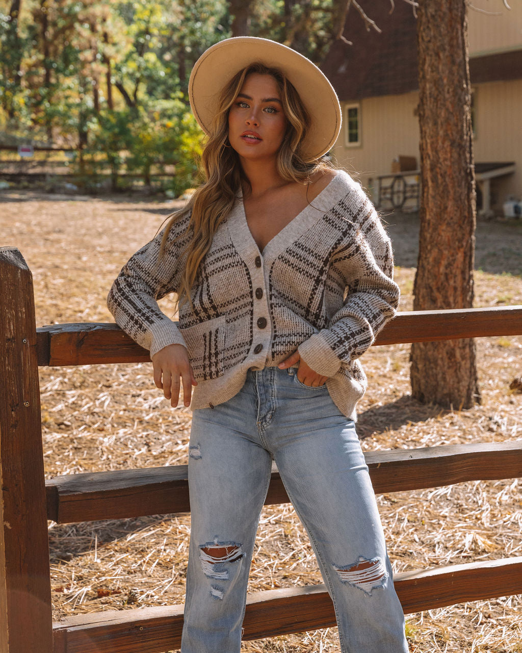Frau in kariertem Strickcardigan und zerrissenen Jeans vor Holzzaun im Herbstwald.