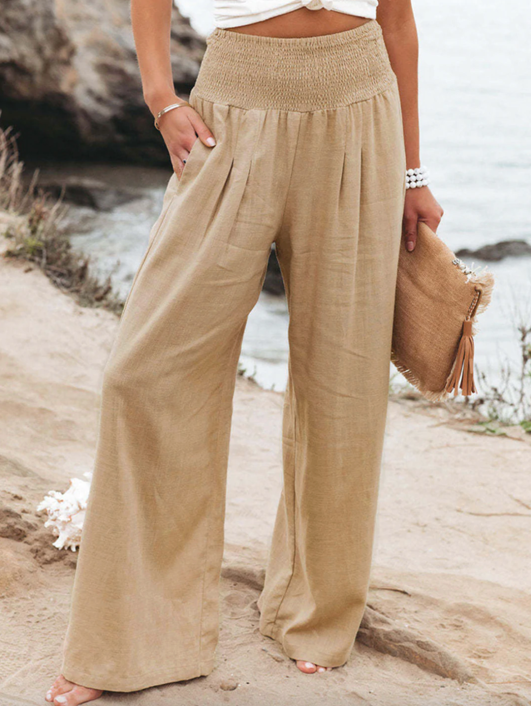 Frau in beigen Leinenhose am Strand, hält eine Strohtasche, Sommermode, lässiger Look.