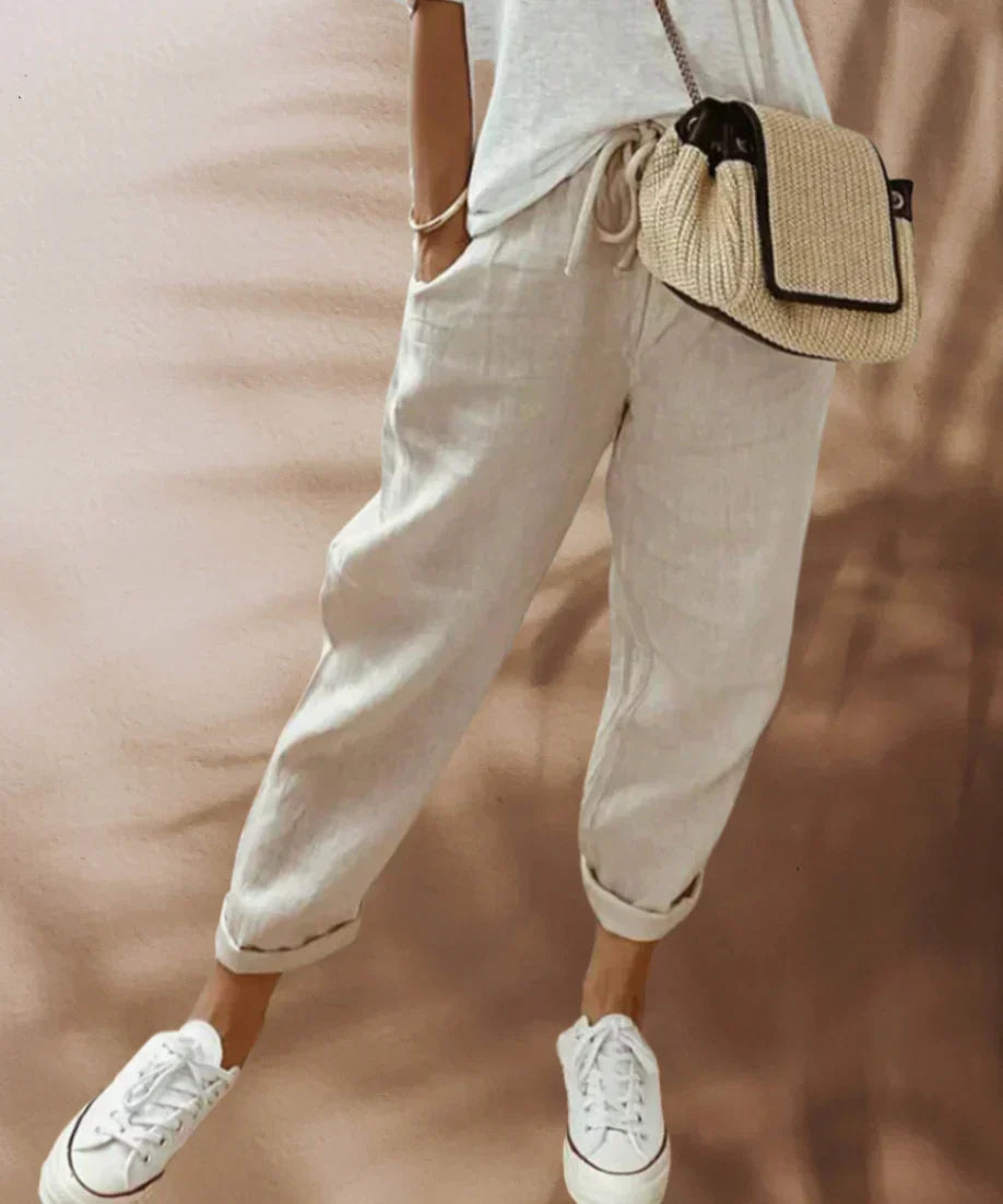 Frau in beigen Leinenhosen, weißem T-Shirt, Strohtasche und weißen Sneakers.