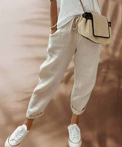 Frau in beigen Leinenhosen, weißem T-Shirt, Strohtasche und weißen Sneakers.