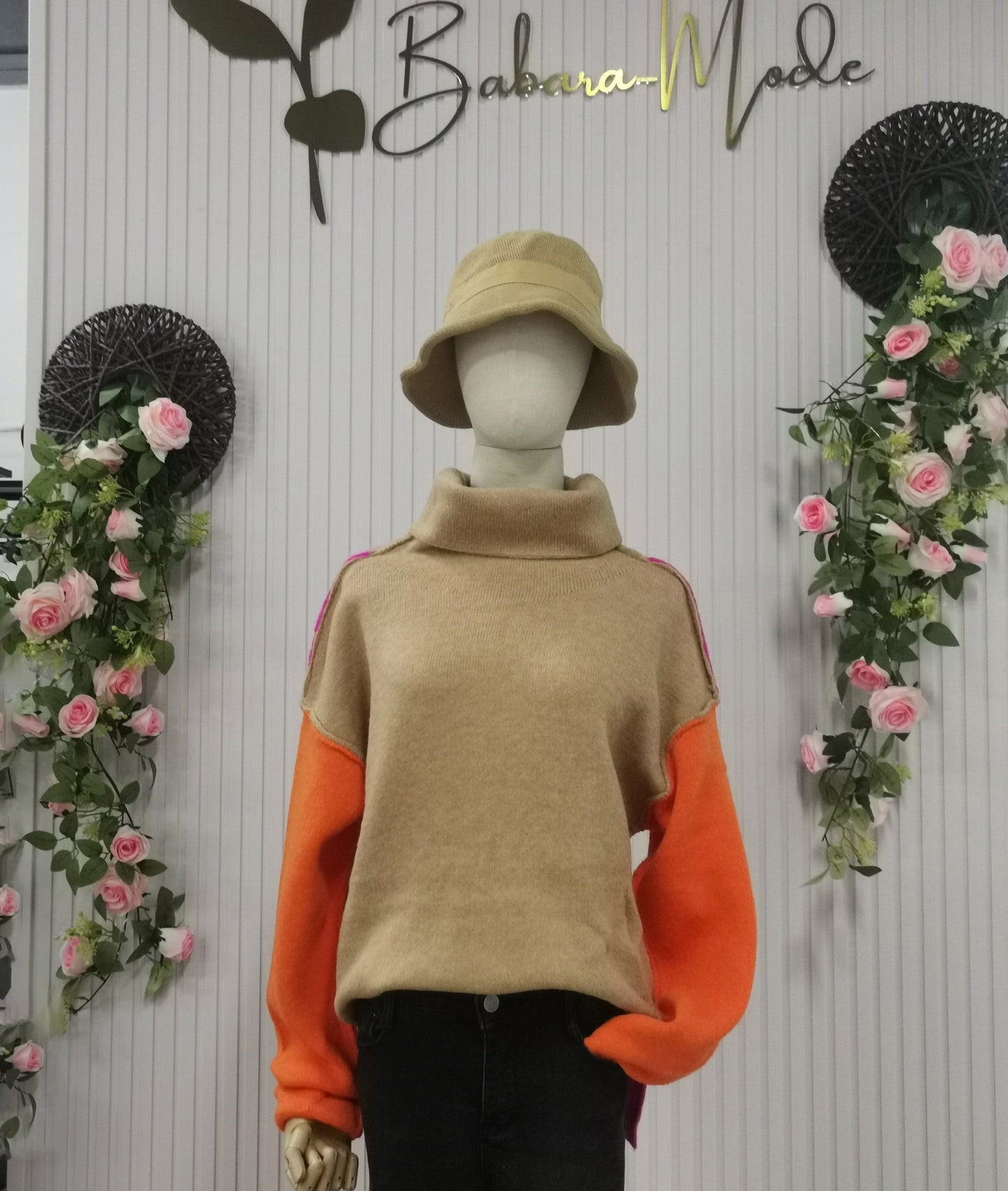 Schaufensterpuppe mit beigem Rollkragenpullover, orangefarbenen Ärmeln, beigem Hut, Blumenhintergrund.