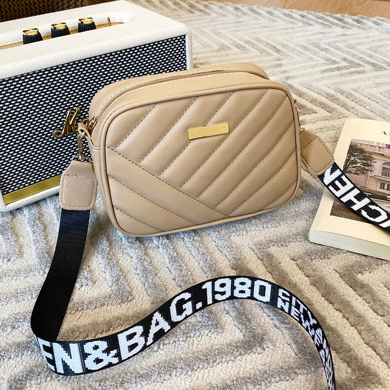Beige Umhängetasche aus Leder mit gestepptem Design und schwarzem Logo-Gurt auf Teppich.