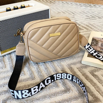 Beige Umhängetasche aus Leder mit gestepptem Design und schwarzem Logo-Gurt auf Teppich.