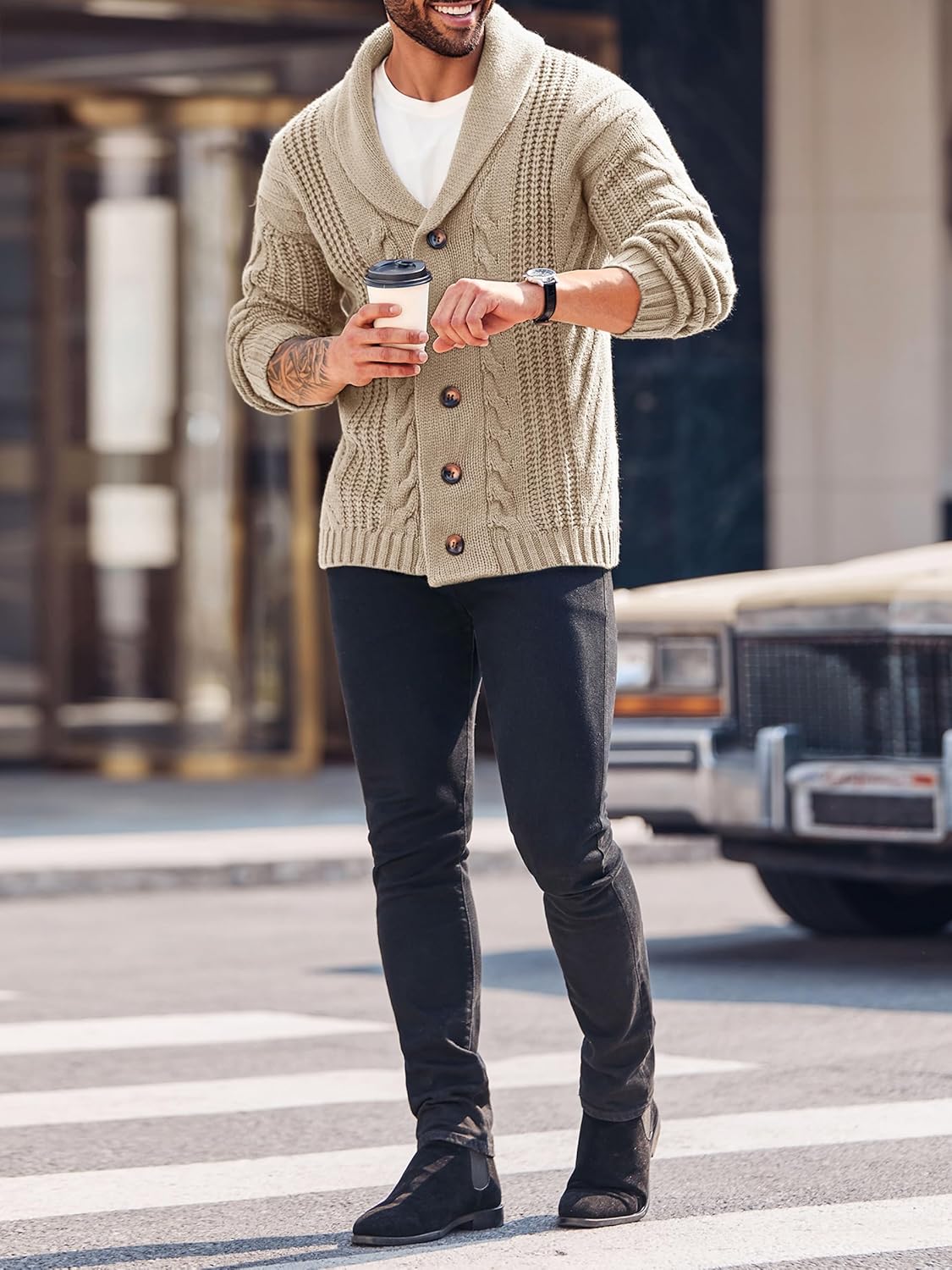Mann in beigem Strickcardigan mit Schalkragen, schwarze Jeans, hält Kaffeebecher, urbaner Hintergrund.