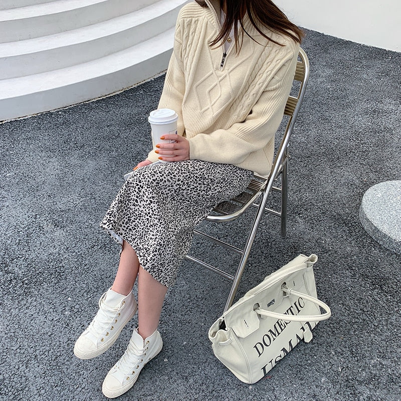 Frau in cremefarbenem Strickpullover, Leopardenrock, weiße Sneakers, hält Kaffeebecher.