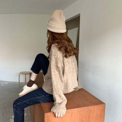 Frau in beigem Strickpullover und Mütze, sitzt auf Holztisch, lässige Wintermode.