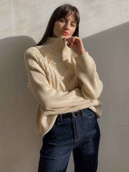 Frau in cremefarbenem Strickpullover mit Zopfmuster, Stehkragen, dunkle Jeans, lässiger Look.