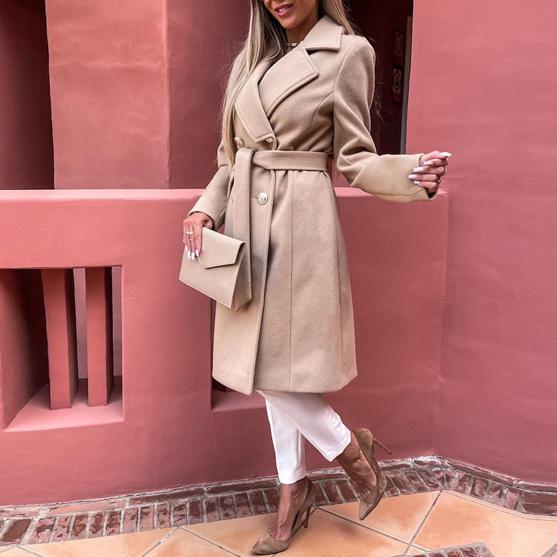 Frau im beigen Wollmantel mit Gürtel, hält Clutch, trägt weiße Hose und High Heels.