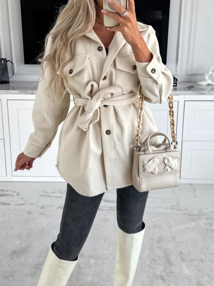 Frau in cremefarbener Jacke mit Gürtel, graue Jeans, weiße Stiefel, beige Handtasche.
