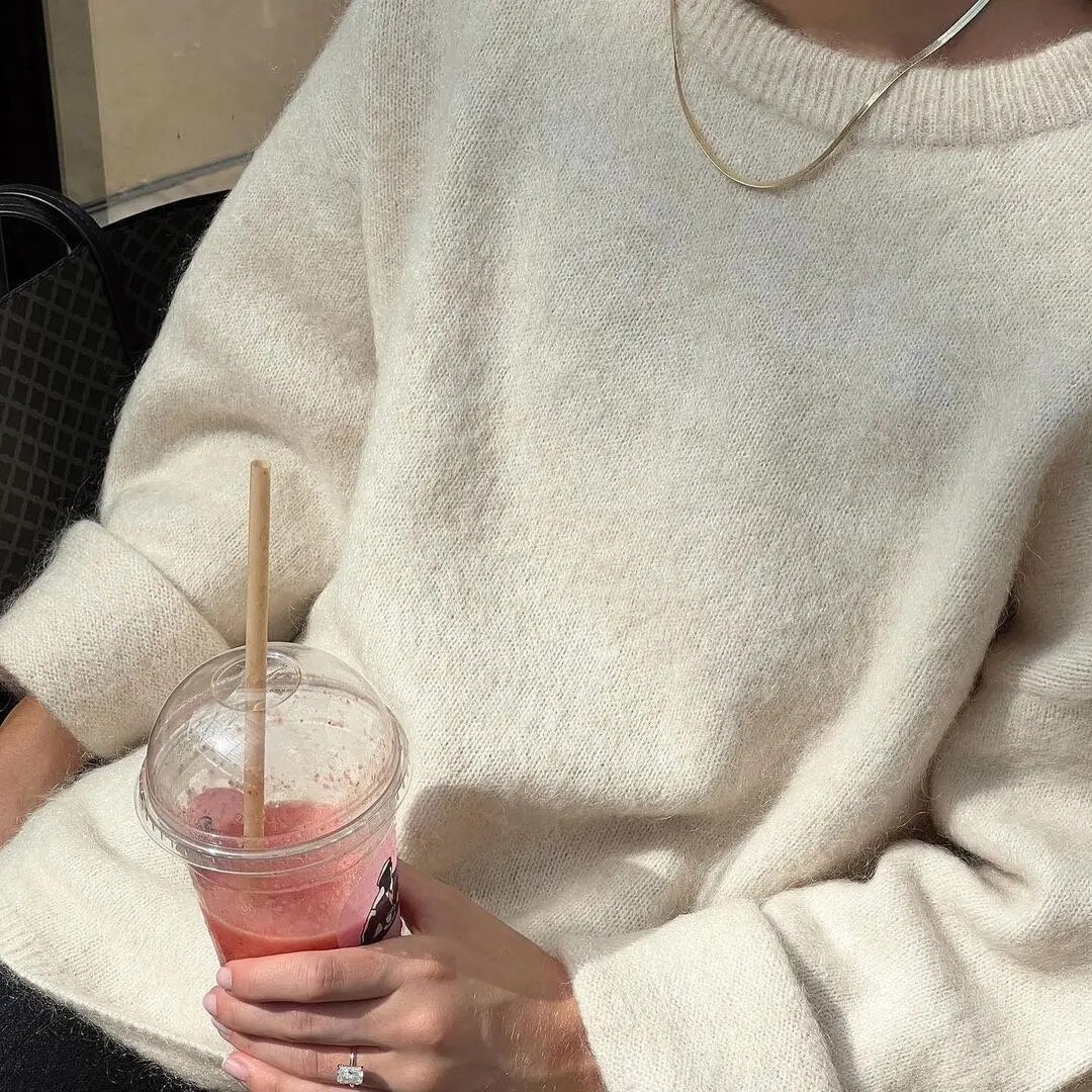 Frau in cremefarbenem Pullover mit Smoothie und Halskette, gemütliche Herbstmode.