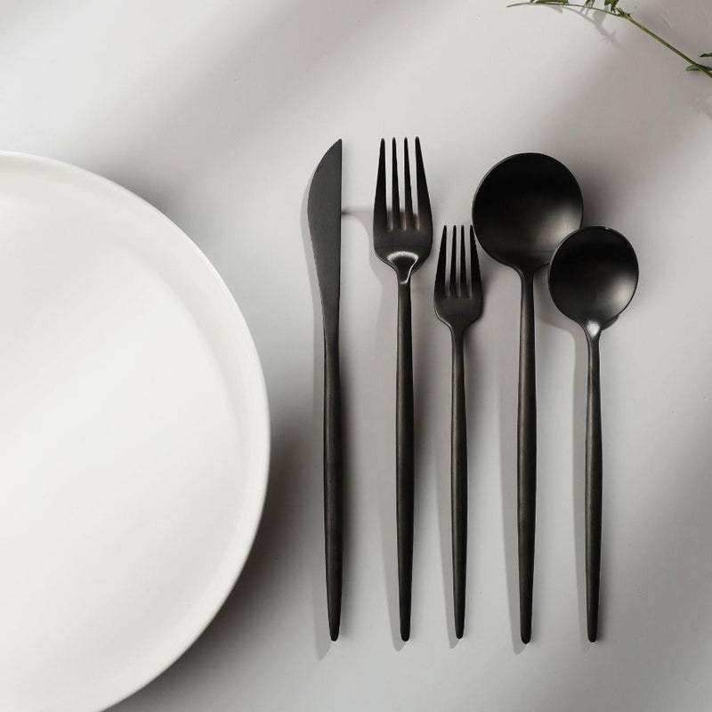 Schwarzes minimalistisches Besteck-Set aus Edelstahl neben weißem Teller auf grauem Hintergrund.