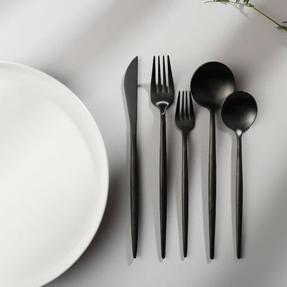 Schwarzes minimalistisches Besteck-Set aus Edelstahl neben weißem Teller auf grauem Hintergrund.