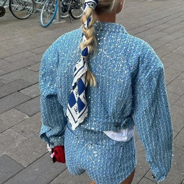 Frau in blauem Pailletten-Outfit mit Zopf und Seidenschal, modisch, urbaner Hintergrund.