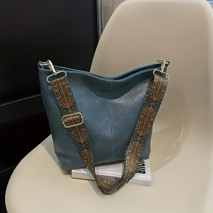 Blauer Ledertasche mit verziertem Schultergurt auf weißem Stuhl, modisches Accessoire.