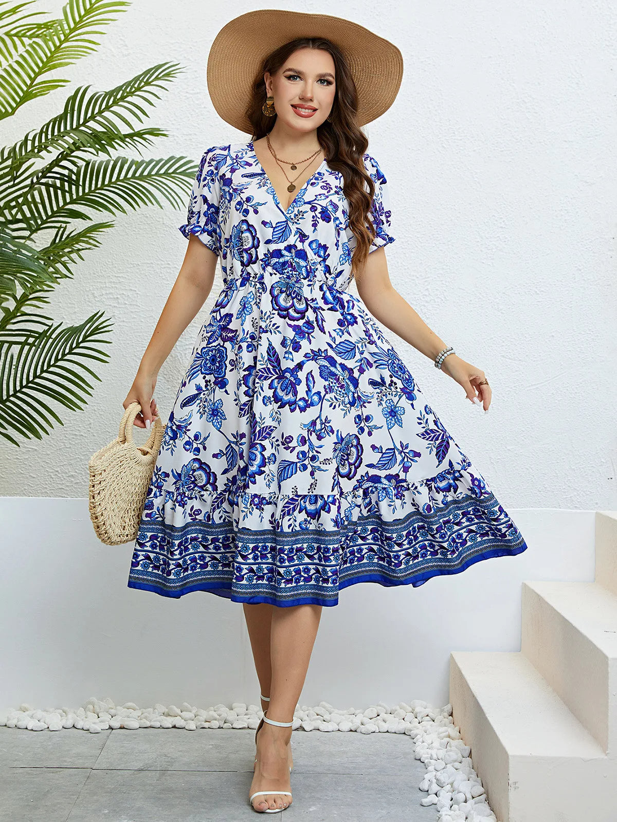 Frau im blauen Blumen-Sommerkleid mit Strohhut und Korbtasche vor weißer Wand.