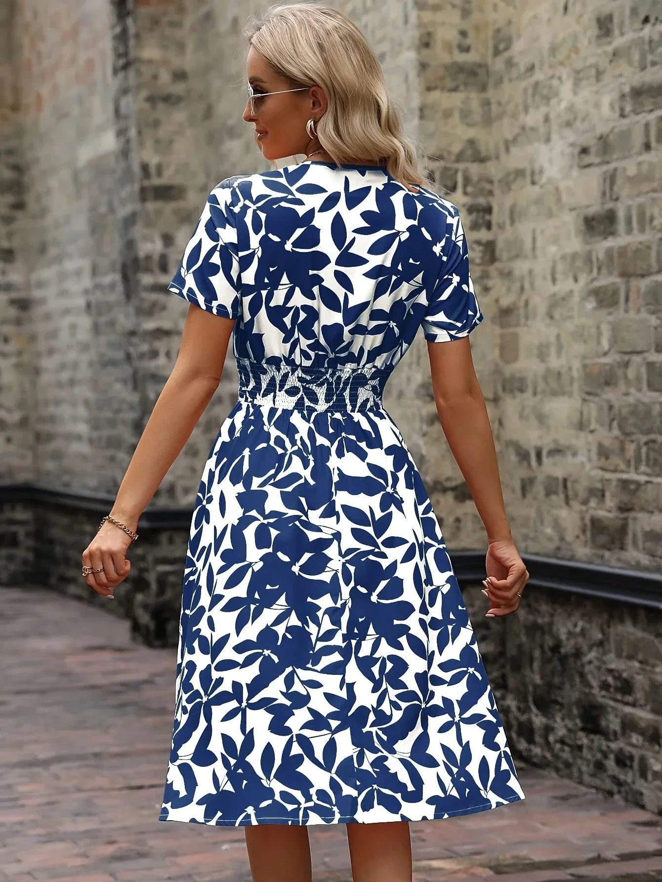 Frau in blau-weißem Sommerkleid mit Blattmuster, steht vor einer Backsteinmauer.