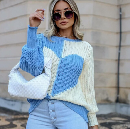 Frau mit blau-weißem Herzpullover, Sonnenbrille und weißer Tasche vor beigem Gebäude.