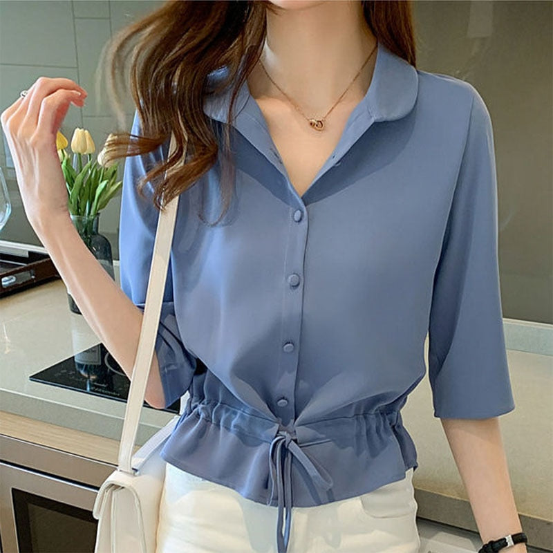 Frau in blauer Bluse mit Knopfleiste und Bindegürtel, weiße Hose, moderne Küche, Tulpen im Hintergrund.
