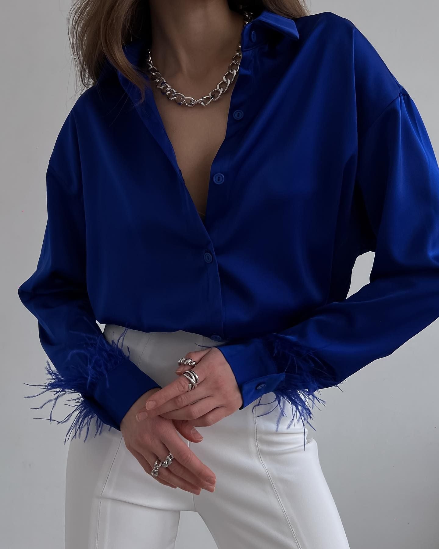 Frau in blauer Satinbluse mit Federn, weiße Hose, silberne Kette, modisches Outfit.