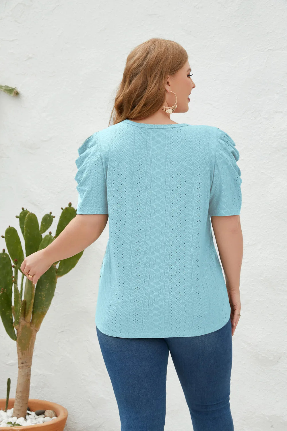 Frau in hellblauer Strickbluse mit Puffärmeln, Jeans, vor weißer Wand, neben Kaktus.