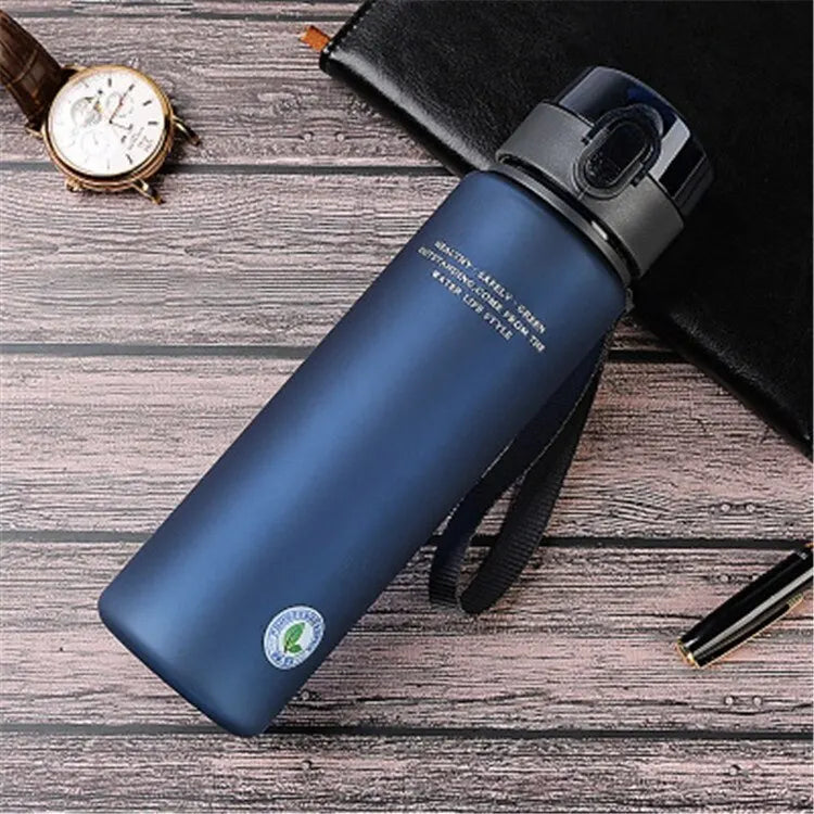 Blaue Edelstahl-Wasserflasche mit Deckel, auf Holztisch, neben Uhr und Stift.