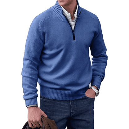 Mann in blauem Pullover mit Reißverschluss, weißem Hemd und Jeans, hält braune Jacke.