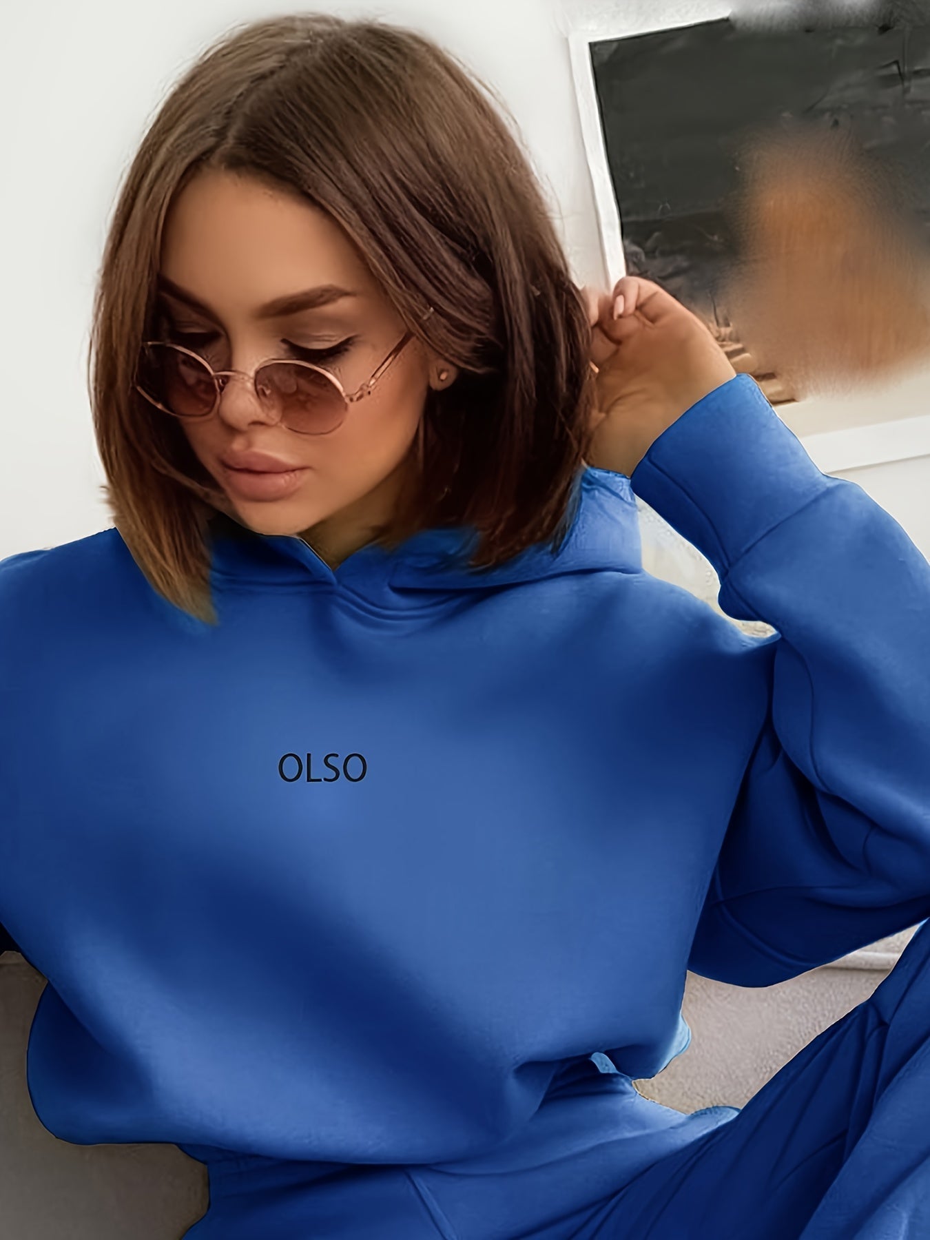 Frau in blauem Olso-Hoodie mit Sonnenbrille, sitzt auf Sofa, modisch, lässiger Stil.