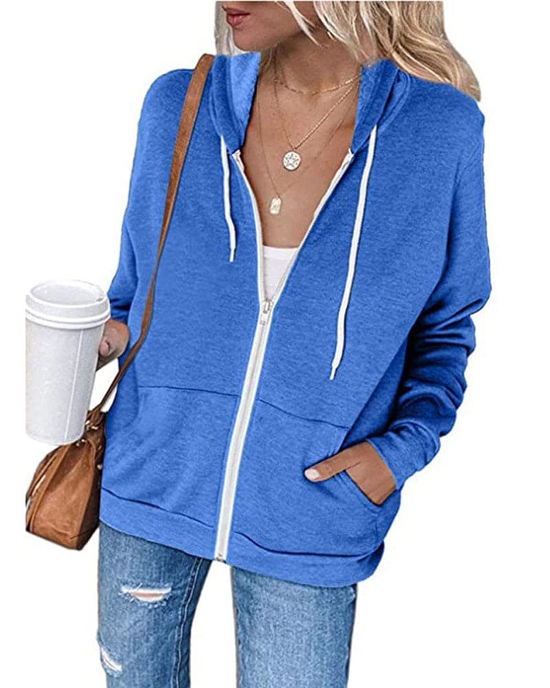 Frau in blauer Kapuzenjacke mit Reißverschluss, hält Kaffeebecher, trägt Jeans und braune Tasche.