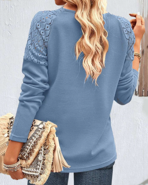 Frau in blauem Strickpullover mit Spitzenärmeln, hält eine beige Fransenhandtasche.