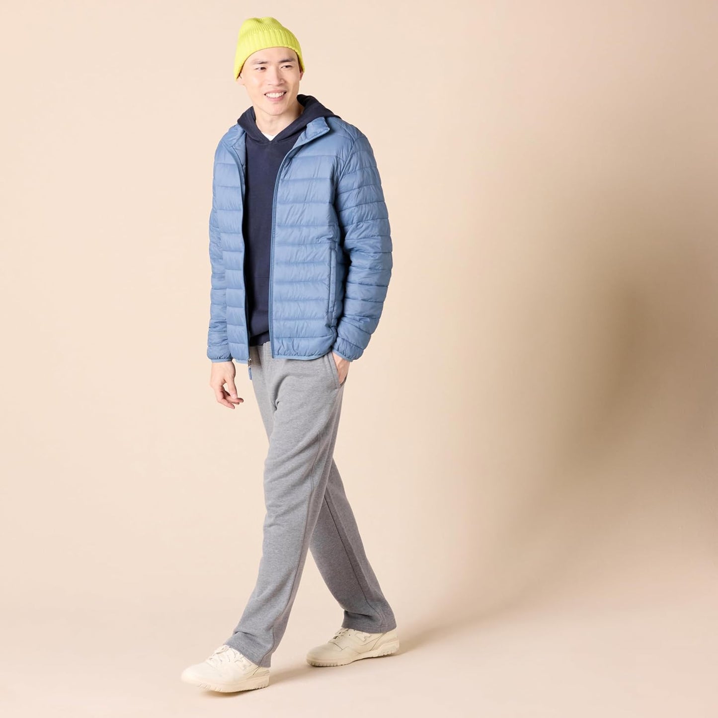 Mann in blauer Steppjacke, grauer Jogginghose und gelber Mütze, lächelnd, beige Hintergrund.