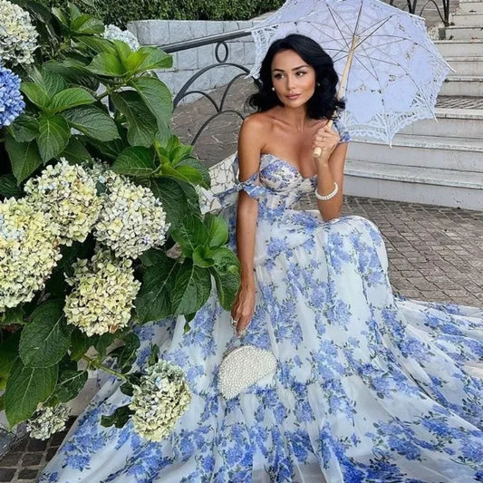 Frau in blau-weißem Blumenkleid mit Spitze, hält weißen Sonnenschirm, sitzt neben Hortensien.
