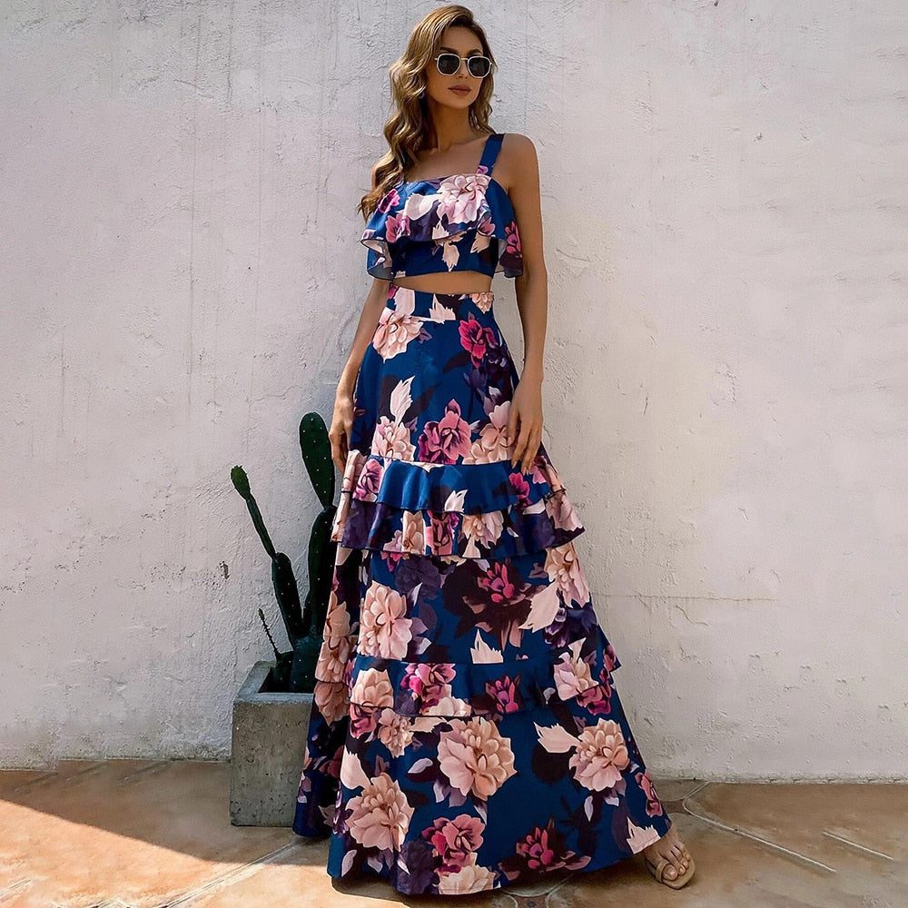 Frau in blauem, geblümtem Maxikleid mit Rüschen, Sonnenbrille, Sandalen, vor weißer Wand.
