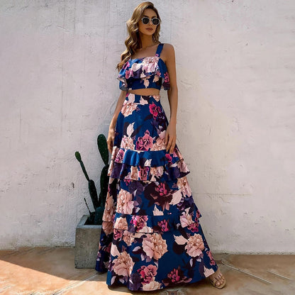 Frau in blauem, geblümtem Maxikleid mit Rüschen, Sonnenbrille, Sandalen, vor weißer Wand.
