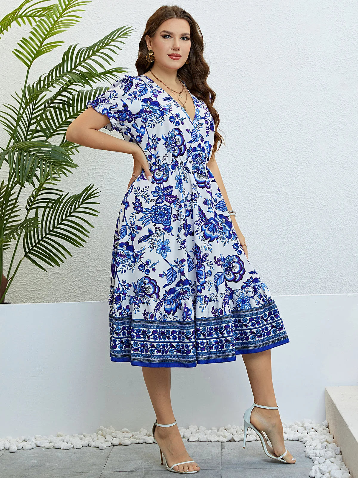 Frau in weißem, geblümtem Sommerkleid mit blauen Mustern, steht vor weißer Wand.