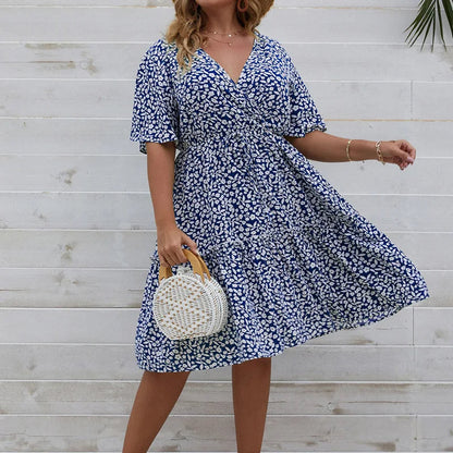 Frau im blauen Sommerkleid mit Blumenmuster, hält runde Strohtasche, vor weißer Holzwand.