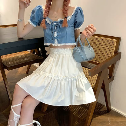Frau in blauem Crop-Top und weißem Rock, hält eine kleine blaue Handtasche, sitzt auf Holzstuhl.