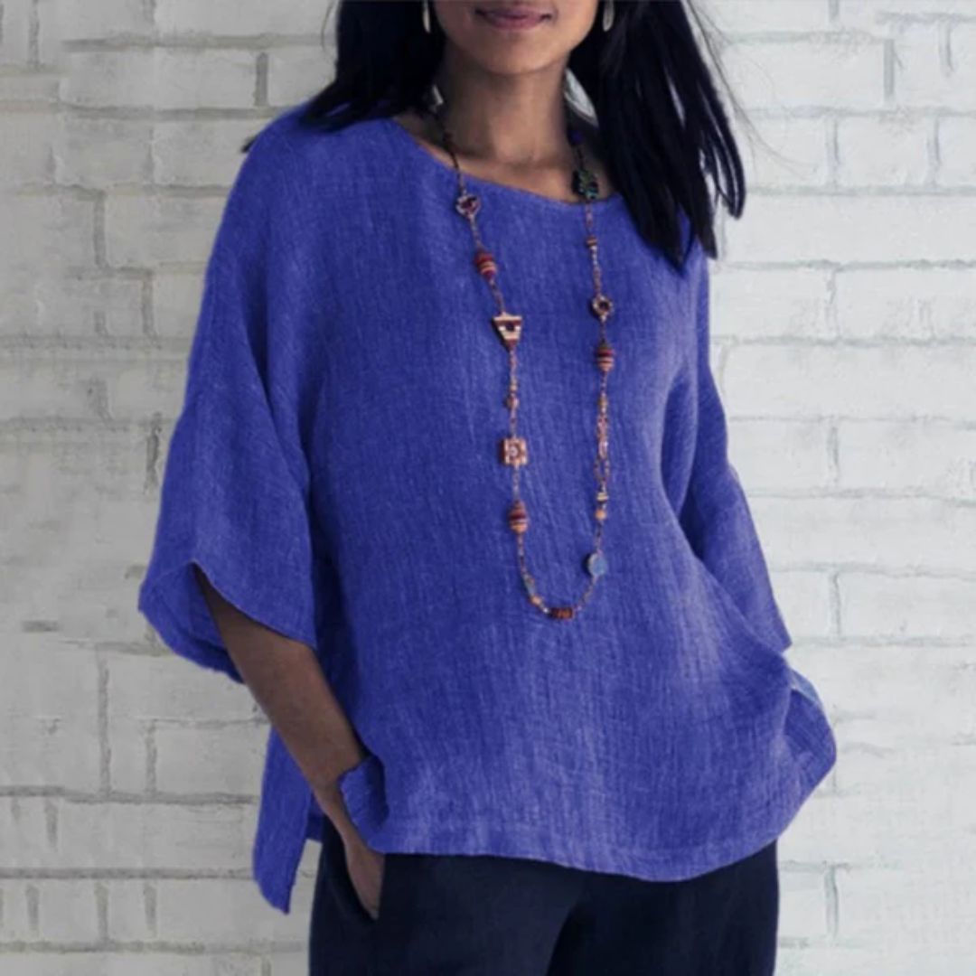 Frau in blauer Leinenbluse mit Holzperlenkette vor weißer Ziegelwand.