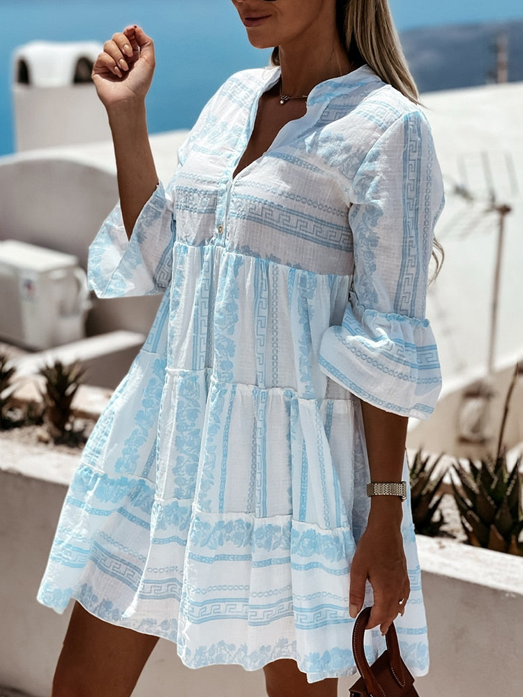 Frau in weiß-blauem Sommerkleid mit Muster, steht draußen, hält braune Sandalen.