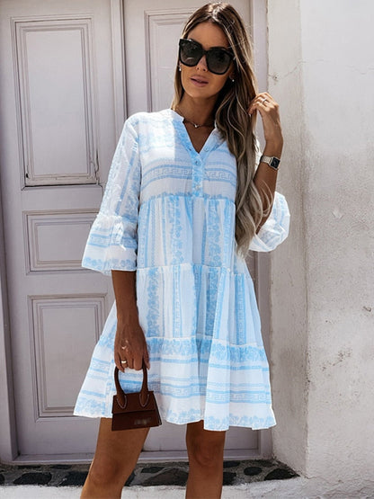 Frau in blau-weißem Sommerkleid mit Sonnenbrille und brauner Handtasche vor Tür.