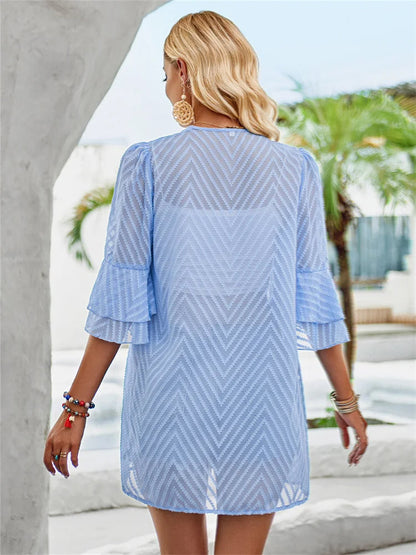 Frau in hellblauem Sommerkleid mit Chevron-Muster, goldenen Ohrringen, Armbändern, Palmen im Hintergrund.