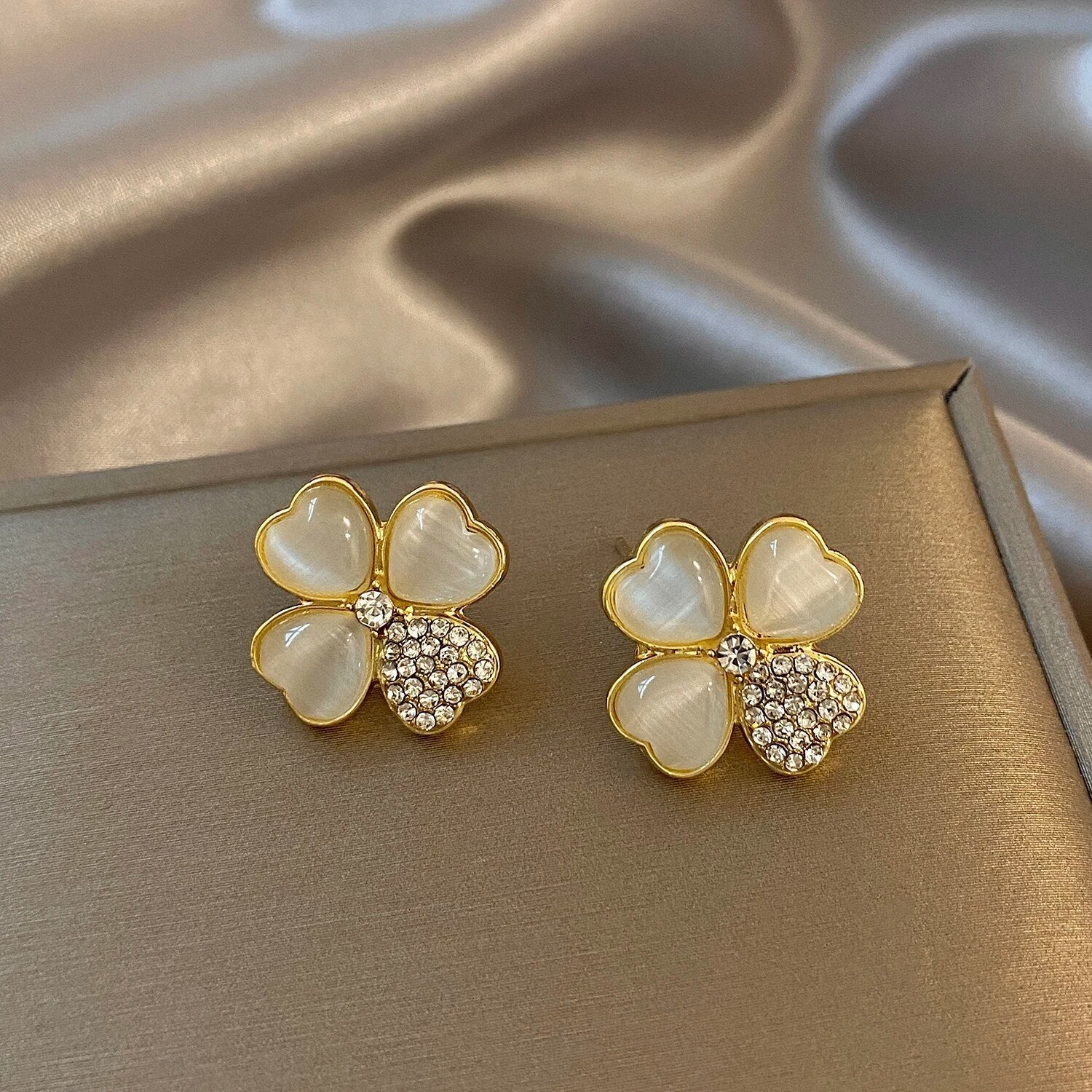 Goldene Ohrringe mit Herzblüten-Design, Perlmutt und Strasssteinen auf Seidenhintergrund.