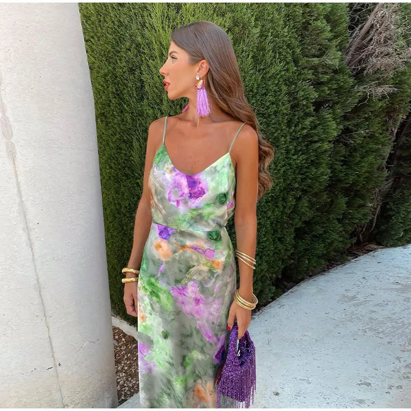 Frau im bunten Sommerkleid mit lila Quastenohrringen und Tasche, vor grüner Hecke.